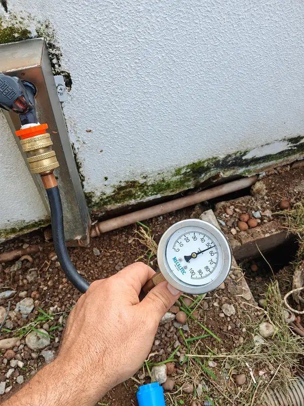 Mão segurando manômetro junto a tubulação e parede.